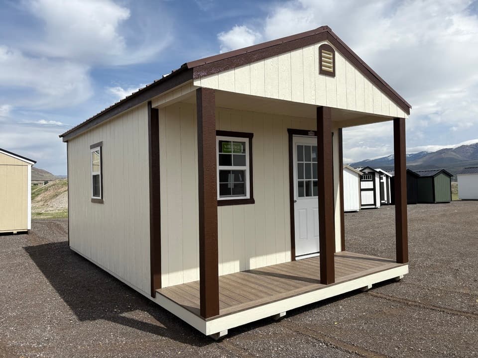 Stor-Mor 12x20 cabin with a covered porch, cream siding and dark brown trim posts, on the JTCS Buildings lot in Burley with the Idaho mountains behind it