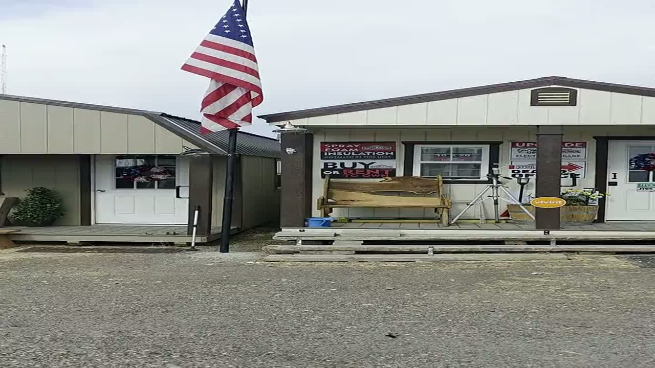 The JTCS Buildings sales lot in Burley, Idaho: the Stor-Mor sales office with an American flag flying out front, a live-edge wood bench on the porch, and buy-or-rent signage in the windows