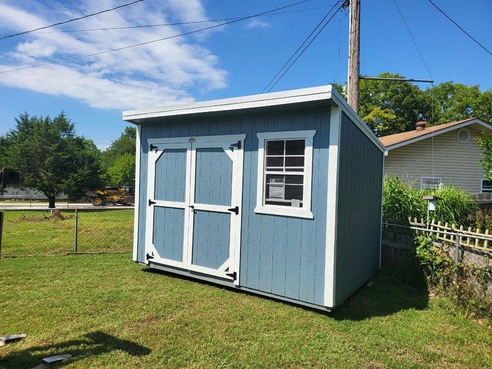 Stor-Mor 8x12 cottage shed in blue-gray with white trim and a single-slope roof, with white-braced double doors and one window, set in a backyard