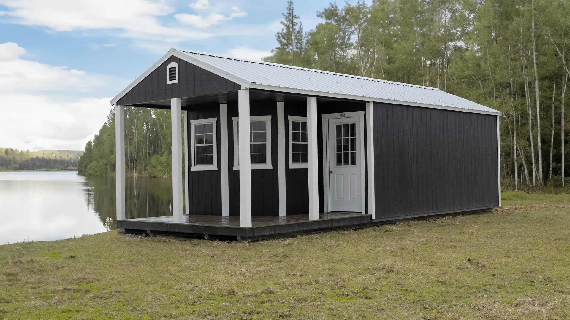 Stor-Mor deluxe cabin in near-black siding with white trim, posts and a white metal gable roof, with a covered porch and three grid windows, set beside a lake