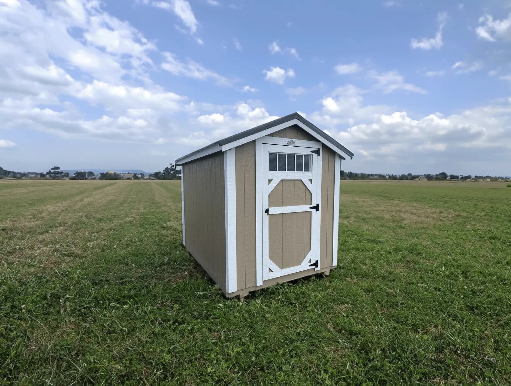 Small Stor-Mor Metro shed in taupe with white trim and a dark gable roof, with a single door topped by a row of transom window panes, set in an open green field