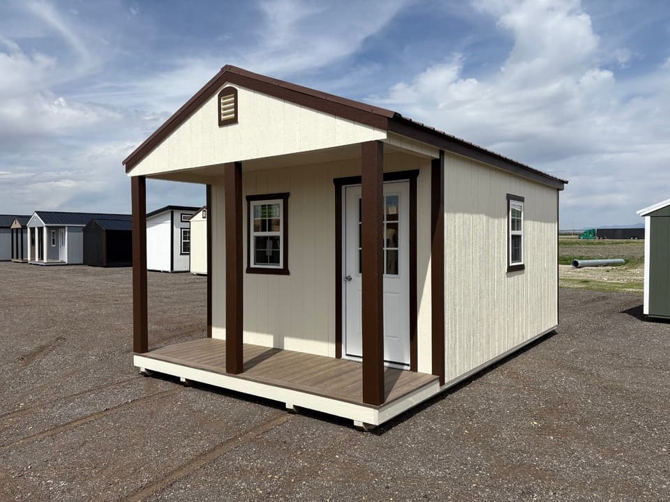 Front corner view of a 12x20 Stor-Mor cabin with a covered porch, white entry door and brown trim, parked on the gravel lot beside a row of storage sheds