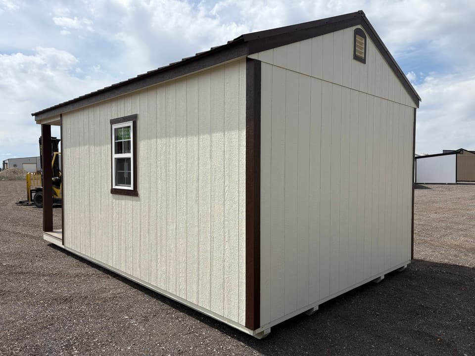 Side view of a 12x20 cabin showing the full length of its siding, a brown-trimmed window and the covered porch, on the JTCS gravel lot