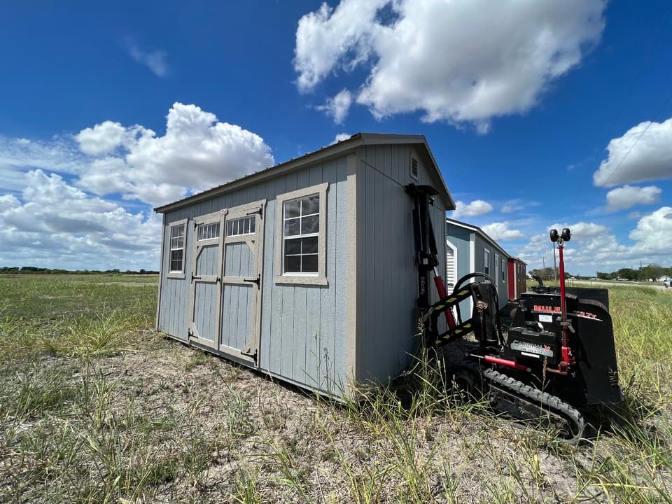A gray storage shed being moved into place on open ground by a red tracked Cardinal shed mover, with more buildings lined up behind it