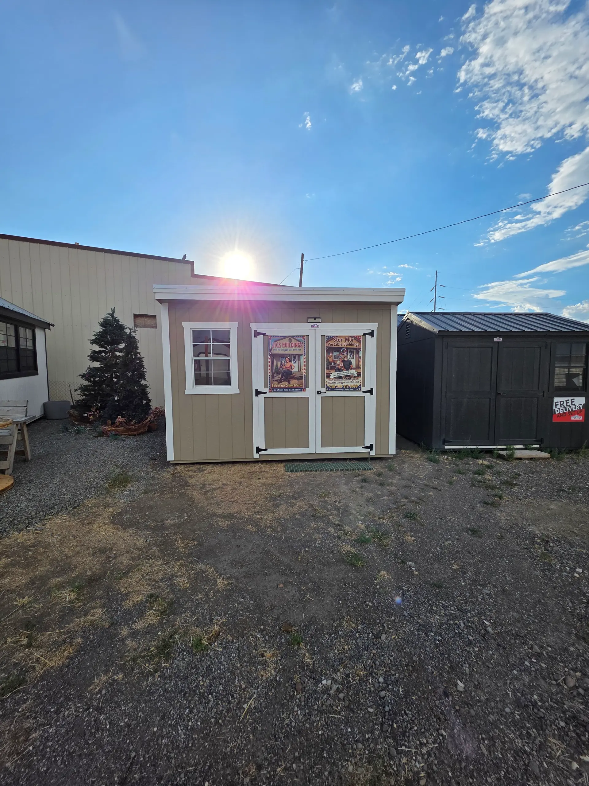 Small taupe garden shed with white-trimmed window and double doors carrying JTCS Buildings posters, backlit by the morning sun on the Burley lot