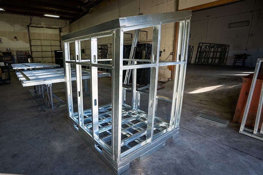 Galvanized steel frame with floor joists and wall studs, sitting on a base inside a workshop with more steel frames in the background