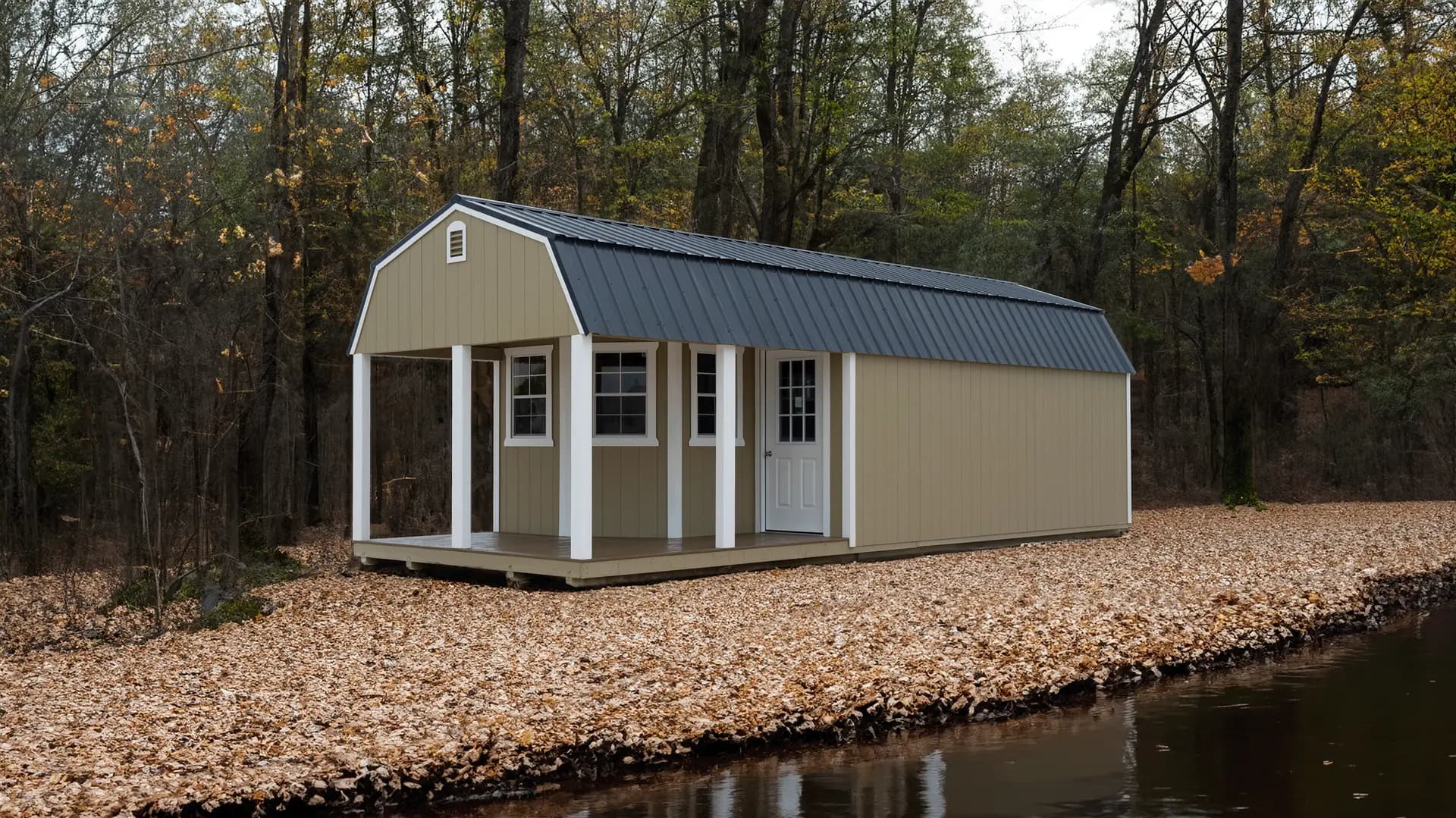 Stor-Mor deluxe lofted barn cabin in khaki with white trim posts and a dark metal gambrel roof, with a covered front porch, set on a leaf-covered riverbank