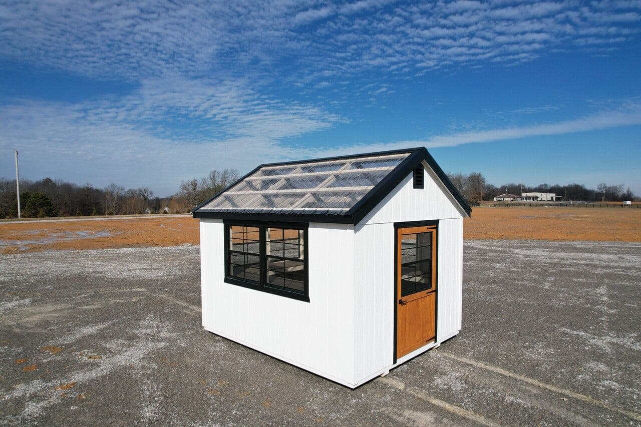 Stor-Mor greenhouse with white siding, black trim, clear polycarbonate roof panels, black-framed windows and a wood-tone entry door, on a gravel lot