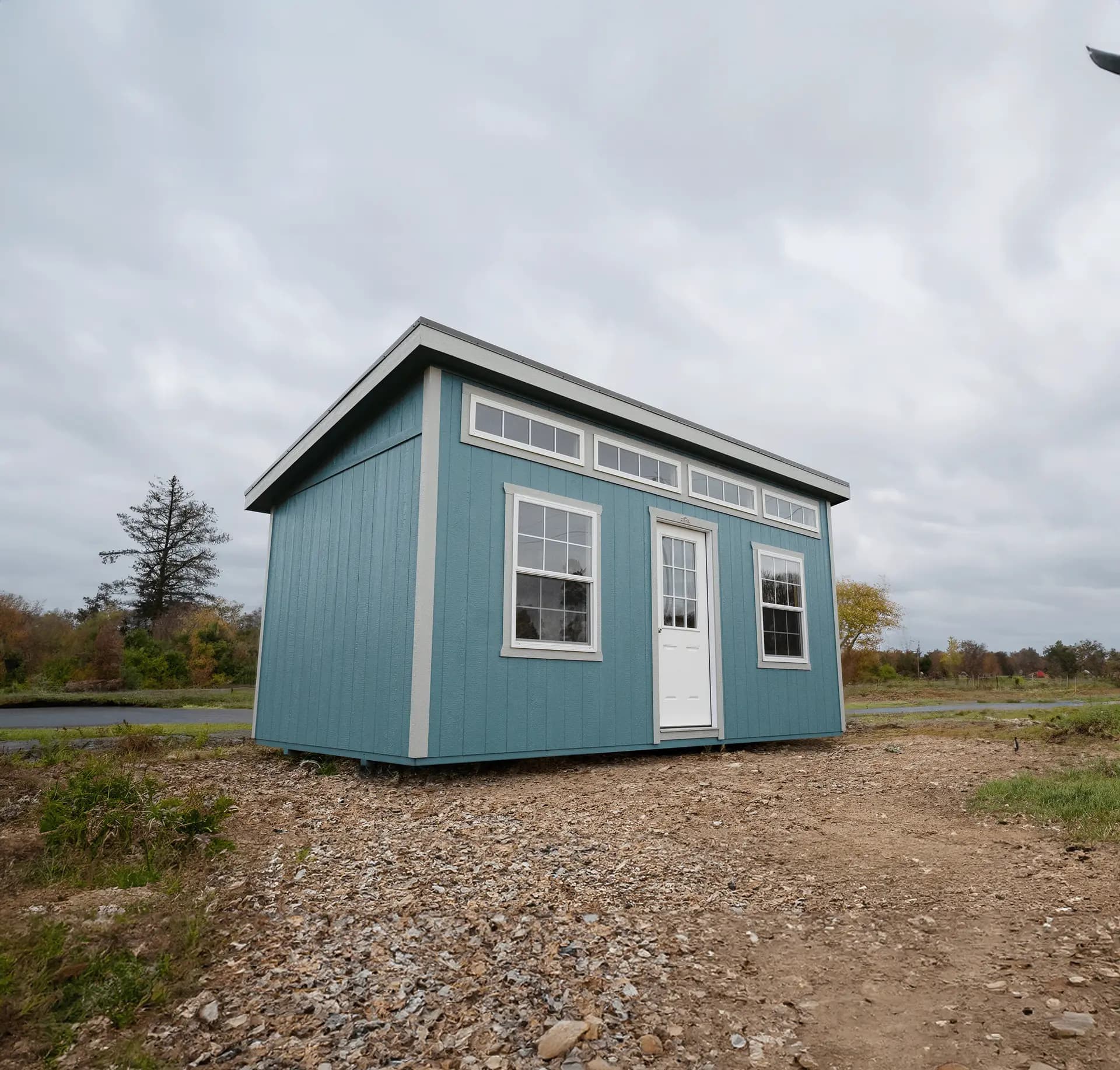 Stor-Mor studio shed in teal blue with cream trim and a single-slope shed roof, with a band of transom windows along the top, a white entry door and two grid windows
