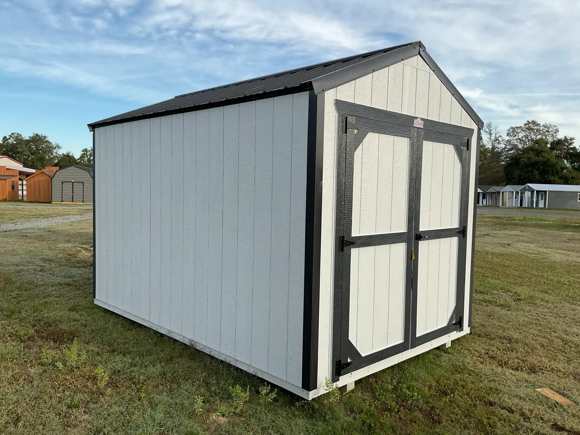 Stor-Mor Tru-Valu utility shed in light gray with charcoal-trimmed double doors and a black metal gable roof, parked on grass with a row of other sheds behind it