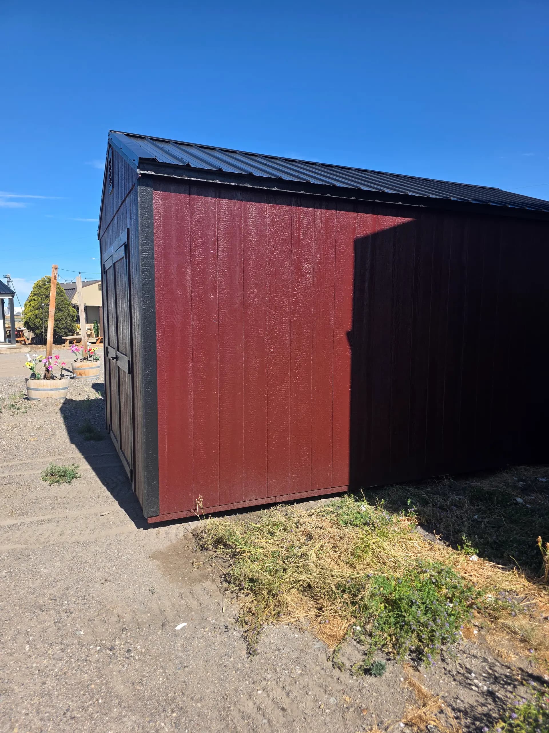 Corner view of a wood-sided storage shed on the Burley lot, painted dark brown with a deep red door wall and a black metal roof
